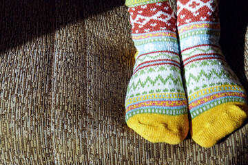 Feet in vibrant multicolored patterned socks with yellow toes resting on a couch. Symbolizes homewear style, Nordic knit tradition, artisan hosiery appreciation, and hygge lifestyle aesthetic.