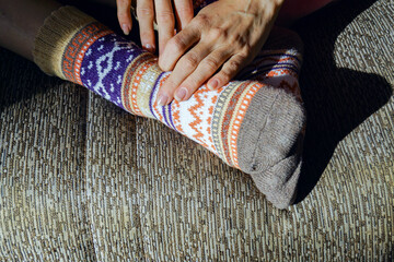A woman’s hands touch her feet wearing woolen socks with an intricate Fair Isle design. Represents traditional knitting craft, cozy home atmosphere, and appreciation of handmade clothing.