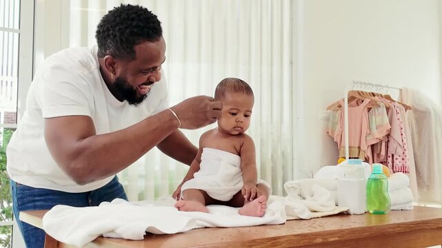 Smiling African dad gently brushes his newborn baby's hair after a bath, showing parenting affection and interaction. Baby care, towels, skincare, and wellbeing in a bright home nursery.