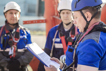 A group of workers in safety gear attending a training session. The instructor reads from a clipboard while others listen attentively. The setting is outdoors.
