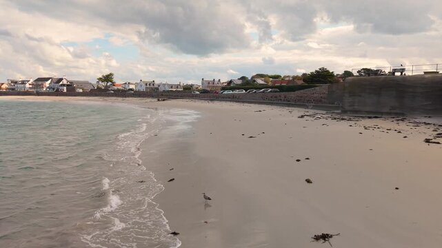 Low drone footage of shoreline at Cobo Bay Guernsey Channel Islands with sandy beach clear water and lapping waves and showing beachfront properties on a bright day with interesting sky