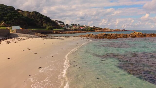 Low drone footage of shoreline at the stunning Cobo Bay Guernsey Channel Islands with golden sand clear turquoise water and lapping waves seabirds and granite rocks on sunny day