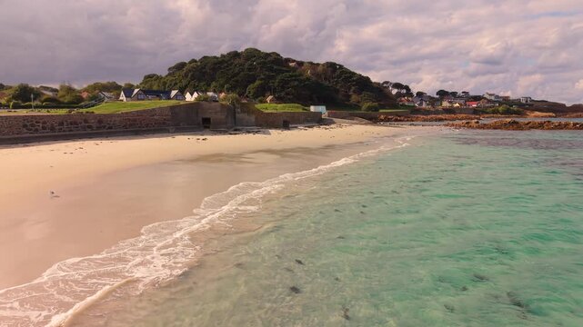 Crystal clear water and golden beach at the stunning Cobo Bay Guernsey Channel Islands with seagull flying by and turquoise water and lapping waves in the sunshine with dramatic sky