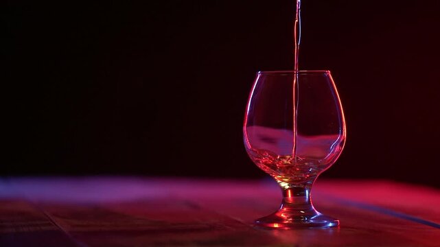 Slow motion footage of pouring cognac, brandy or whiskey into an empty clear glass standing on a wooden tabletop.