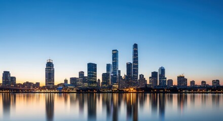 Obraz premium Panoramic view of the illuminated mumbai skyline at dusk reflecting in the calm water of the arabian sea with a clear blue sky