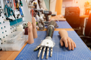Various artificial hands and a partial arm rest on a gridded workbench in a well-lit workshop. Tools hang above the work surface used to create and customize prosthetics