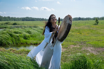 A beautiful female shaman in a trance in the white dress drumming in the natural environment