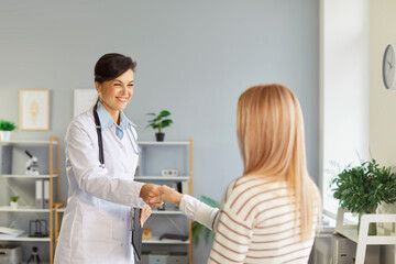 Obraz premium Portrait of female smiling young friendly doctor shaking hands with woman greeting her patient standing in medical clinic. Physician working in office. Medicine and health care concept.