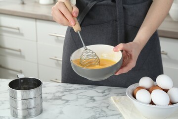 Woman beating eggs with whisk at white marble table in kitchen, closeup