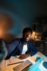 Vertical selfie of happy young African American man looking at camera while studying late at night at home office.