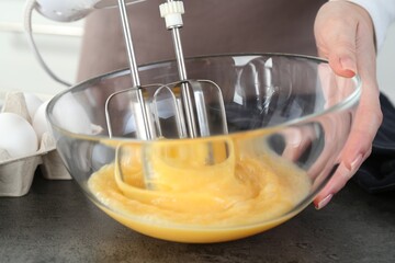 Woman beating eggs with mixer at grey table in kitchen, closeup