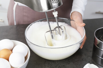 Woman beating egg whites with mixer at grey table in kitchen, closeup