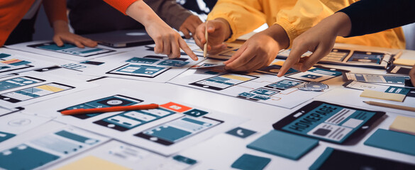 A diverse group of individuals collaborates on user interface mockups at a table filled with notes and tools, emphasizing teamwork in design and innovation. SACTR