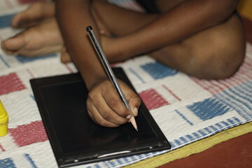 A young Indian boy sits on a woven mat, deeply focused as he writes on a digital writing pad,...