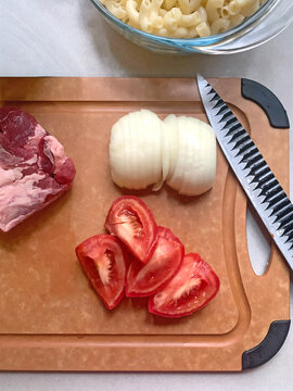 The raw macaroni, steak beef, oinion, tomato, knife and wooden chopboard