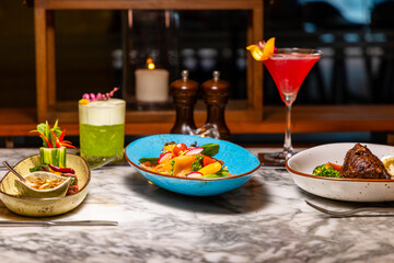 Gourmet dinner setting with colorful salads, cocktails, marble table, and elegant lighting in upscale restaurant.