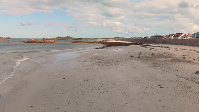 Low forward drone footage of shoreline at Cobo Bay Guernsey Channel Islands with deserted sandy beach and lapping waves with rocky outcrops on a bright day with interesting sky