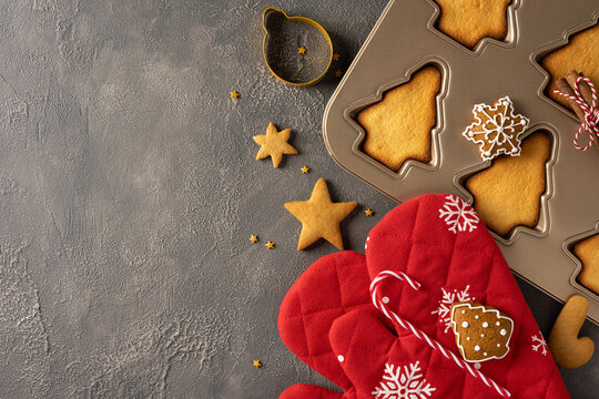 Seasonal baking setup showing a metal cookie sheet with tree shaped cookies, star cookies, a candy cane and a red quilted oven mitt, evoking Christmas treats and New Year celebrations