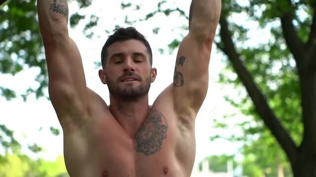 A shirtless man with tattoos is performing a pull-up exercise outdoors in a park with trees in the background.