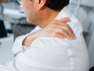 Close-up of Asian Businessman hand massaging his neck, shoulder suffers from pain while working on computer overworking at night in office. Male manager office syndrome, muscle pain from working.