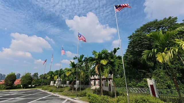 Five flagpoles stand tall, flying the Malaysian flag proudly against the midday sky. The flags gently wave, representing national identity and unity under the bright sun.