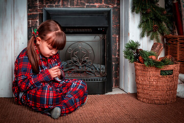 Happy little girl child in pajamas sitting in christmas decorated room near fireplace and prays. Christmas. New Year. St. Nicholas Day. Cozy, festive bedroom interior. Make a wish.