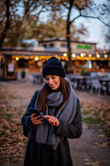 Stylish middle-aged woman wearing long coat and knitted beanie and using smartphone outdoors