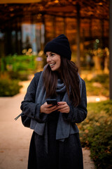 Happy woman wearing beanie and knitted scarf and using her smartphone on the street