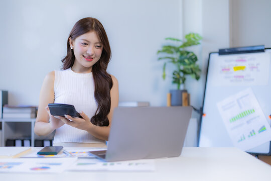 Asian businesswoman working on financial data at her office desk, calculating numbers with a calculator and using a laptop, representing business management and accounting
