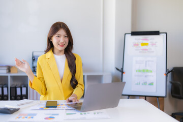 Asian businesswoman working on laptop, presenting business ideas and cheerful, sitting at office desk with documents, and whiteboard showing charts in background
