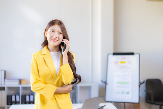Young Asian businesswoman smiling, standing in a contemporary office setting while making a professional phone call, reflecting successful communication and business interaction