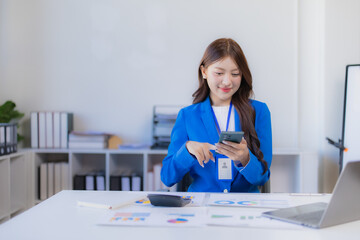 Smiling Asian businesswoman in a blue blazer uses smartphone at a desk with laptop, charts and calculator, portraying modern digital communication, finance and workplace productivity