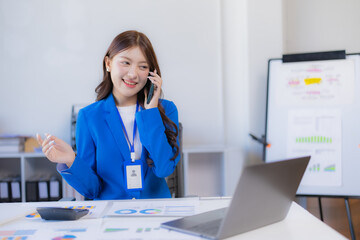 Asian businesswoman in a blue blazer smiling and talking on a mobile phone, holding an employee ID card, with a laptop and financial documents on the desk in a bright office environment