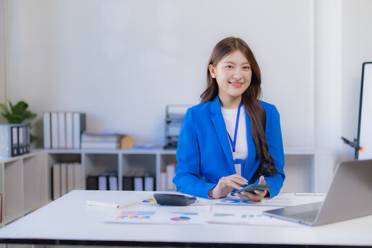 Asian businesswoman working at her desk, calculating financial data using a smartphone and calculator while looking at the camera with a confident smile in her blue suit