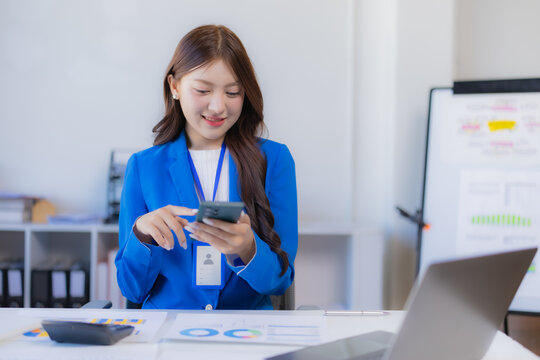 Asian businesswoman working at desk, smiling while using a mobile phone, representing modern communication, business productivity, and digital finance in a professional office environment