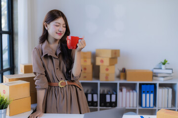 Young Asian businesswoman relaxing with a red mug in her home office, managing an e-commerce store surrounded by shipping boxes, laptop, and packaging supplies, smiling contentedly