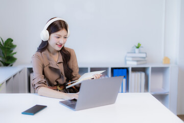 Young Asian woman wearing headphones while looking at a laptop and writing in a notebook, actively engaged in online education, remote work, or virtual classroom activities from a home office