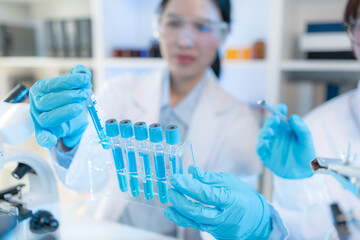 Fototapeta premium Two female scientists in white lab coats working together in a bright laboratory with microscopes, glassware, and blue test tubes, representing teamwork and modern scientific research environment.