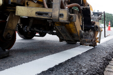 Worker operating road marking machine applying white line on new asphalt pavement. Road maintenance and construction concept, ensuring traffic safety and infrastructure quality.