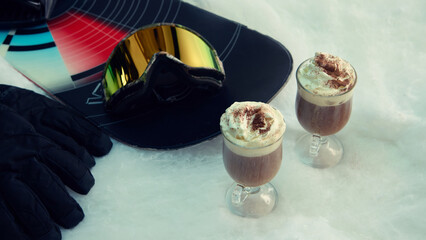 Two hot drinks with whipped cream and snowboard gear on snow surface. Concept of winter tourism marketing, lifestyle media, apres-ski energy, and emotional leisure promotion.