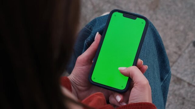 Woman holding smartphone with green screen mockup outdoor, sitting on the bench at the street using phone for chatting with friends, surfing social media, watching reels, video, reading book during re