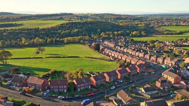 Sweeping aerial pan of Hemingfield village showing rows of brick houses, curved streets, green fields, and forested hills under soft afternoon light in South Yorkshire.