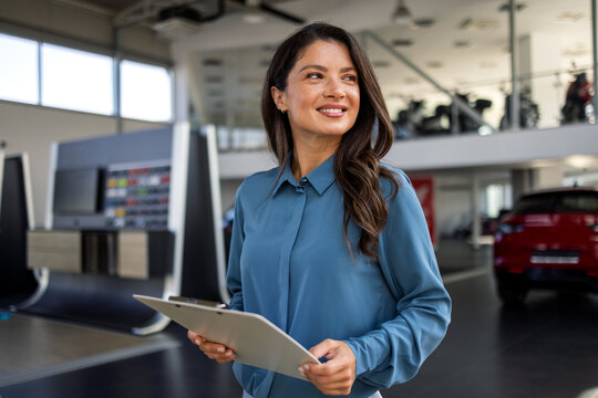 Confident female car sales agent in business attire holding a clipboard, standing beside a sleek new vehicle inside a bright dealership showroom.