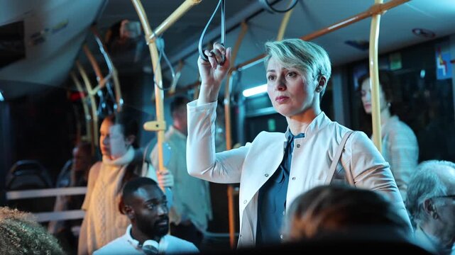 Exhausted businesswoman with short blonde hair standing in a crowded bus, holding onto a strap hanger while commuting home from work late at night, surrounded by other diverse passengers