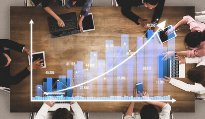 A business team collaborates during a meeting, analyzing digital graphs on laptops and tablets, discussing strategies for growth and performance improvement. Tessel