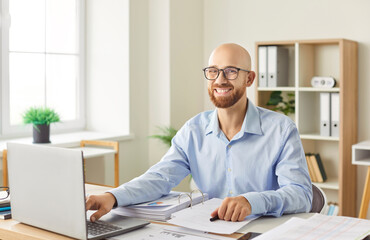 Portrait of a happy, bald businessman working with laptop and documents in the office. Success and...