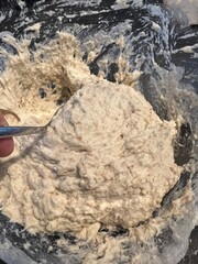 Thick, sticky bread dough with visible grains being mixed in a glass bowl using a metal spoon