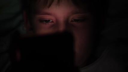 A close-up of a Caucasian teenager's face illuminated by the light of a gadget's screen in the dark, a child using a smartphone in the dark in bed - Powered by Adobe
