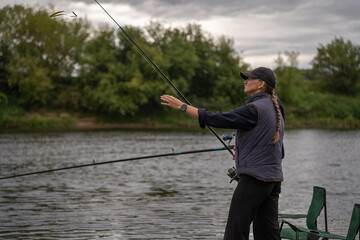 Fisherwoman wearing a cap and vest is casting her fishing rod into a river on a cloudy day, surrounded by vibrant green vegetation and enjoying the tranquility of nature