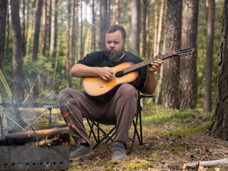 Bearded man is playing an acoustic guitar while sitting on a portable chair in a forest next to a campfire and a tent, enjoying a peaceful camping trip